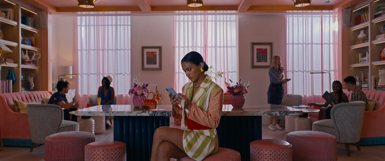 Do Revenge (2022) movie still: medium — A stylish, pastel-colored cafe interior features large windows draped in sheer pink curtains. A woman in a…
