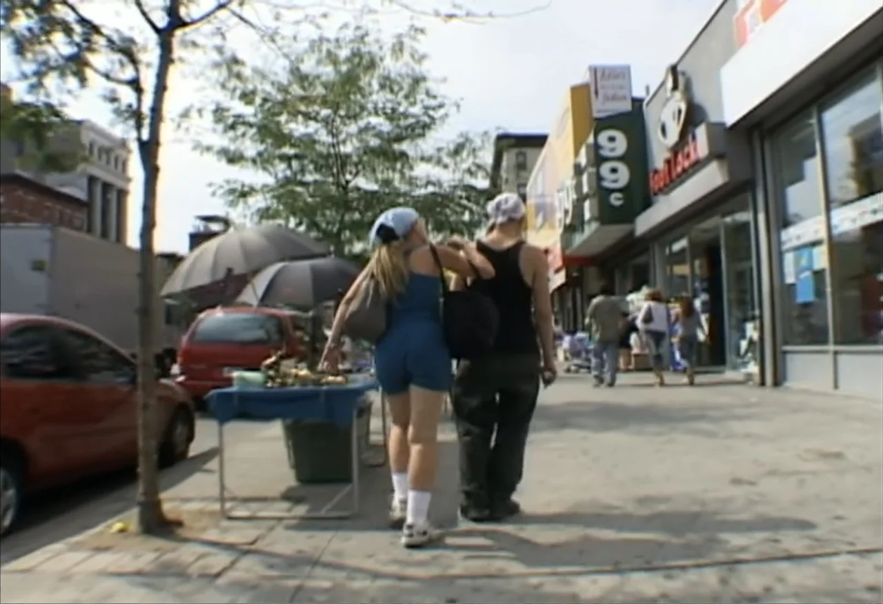 Dope Sick Love (2005) movie still: fullbody — A sunny urban commercial street with storefronts and a small sidewalk market stand. Two young women…