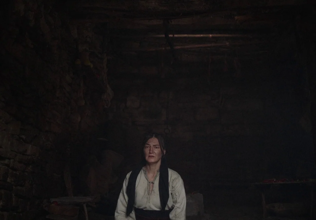 You Won’t Be Alone (2022) movie still: medium — A dimly lit room features rough stone walls and low timber rafters above. A woman sits in the…