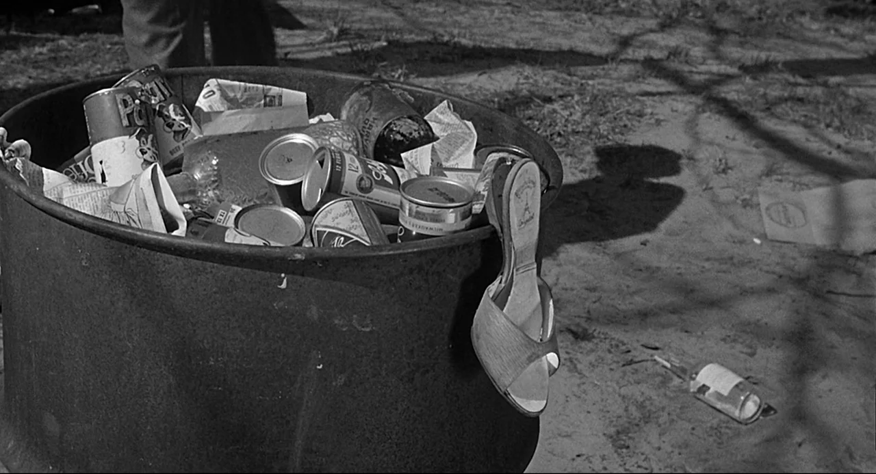 Anatomy of a Murder (1959) movie still: closeup — A metal drum filled with discarded metal cans sits on a patch of dirt. A lone high-heeled sandal…