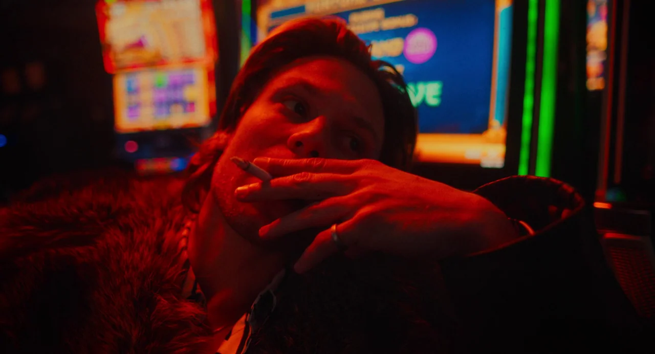 My Animal (2023) movie still: closeup — A dimly lit area of a casino floor is crowded with colorful glowing slot machines. A young man wearing a…