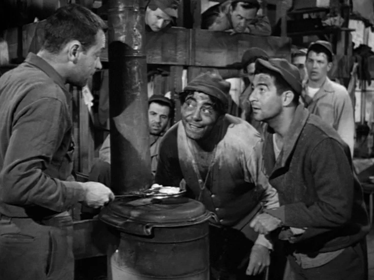 Stalag 17 (1953) movie still: medium — A dimly lit barracks room filled with metal bunk beds and wooden structures. Several men gather around a…