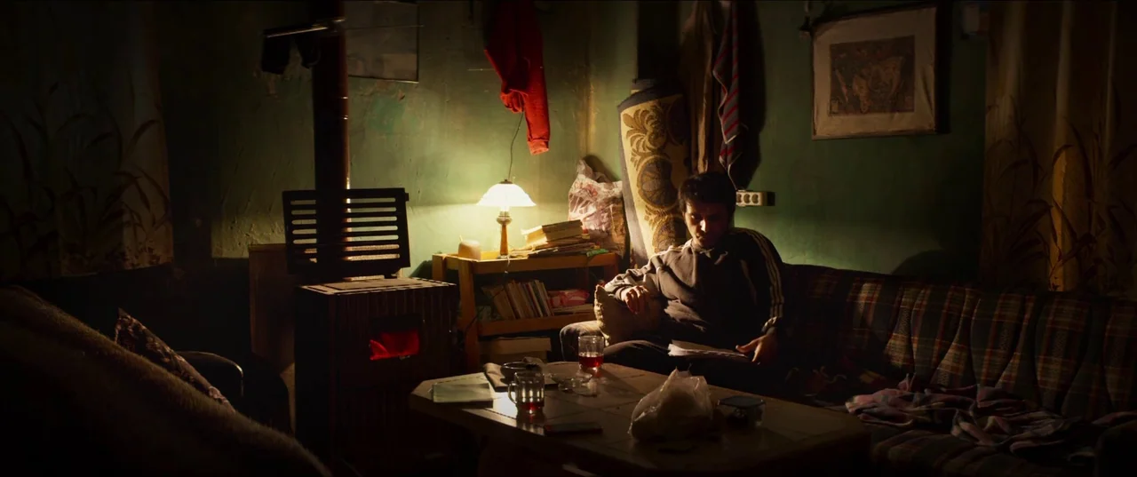 About Dry Grasses (2023) movie still: medium — A dimly lit room features olive-green walls decorated with a single framed print. A young man sits…