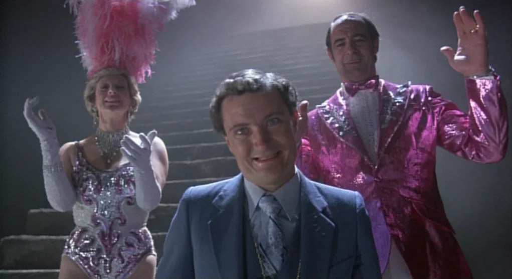 Time Bandits (1981) movie still: medium — Three performers stand on a staircase inside a dark, open performance space. The group smiles and gestures…