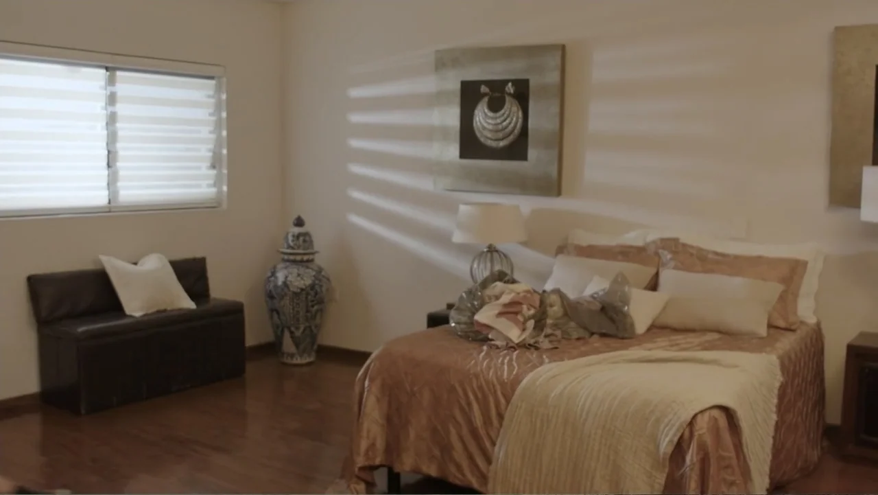 Por Amor al Money (2023) movie still: medium — A well-lit bedroom features a bed with beige bedding and a large ornate vase. The room is quiet with…