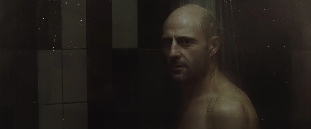 Anna (2013) movie still: medium — A modern shower stall features dark, rectangular wall tiles and constant overhead water flow. A bald man stands…