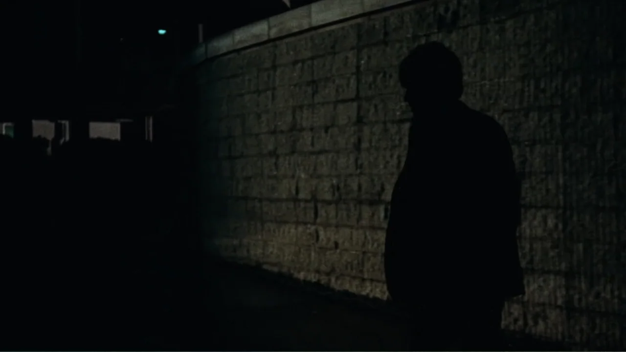 All the President's Men (1976) movie still: medium — A dim exterior parking garage features a textured light-colored brick wall at night. A lone man…