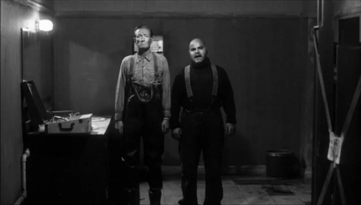 The American Astronaut (2001) movie still: fullbody — A dark utility room features plain walls and a tiled floor. Two men in work attire stand side…