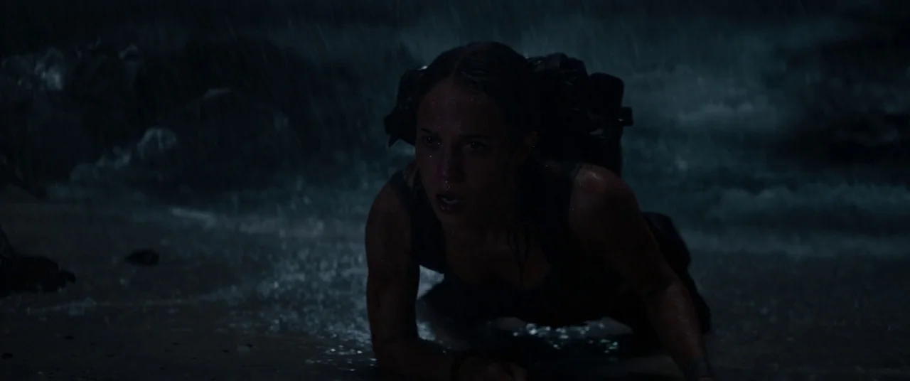 Tomb Raider (2018) movie still: medium — A dark, storm-swept beach at night with rain falling against the sand. A woman crawls along the wet…