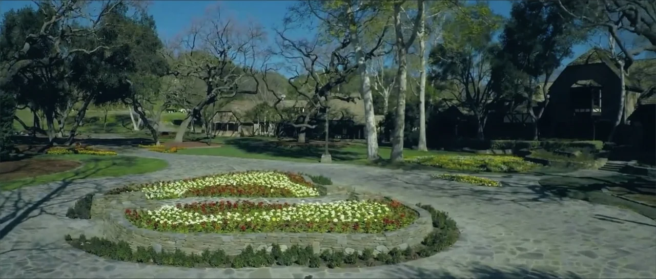 Square One (2019) movie still: establishing — A stone-paved park area features a circular flowerbed with red and white blooms. Mature trees with…