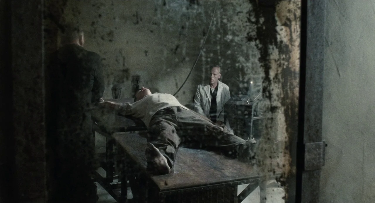 1984 (1984) movie still: wide — A dark, industrial laboratory room features peeling paint and a heavy, cluttered glass partition. A man lies on a…
