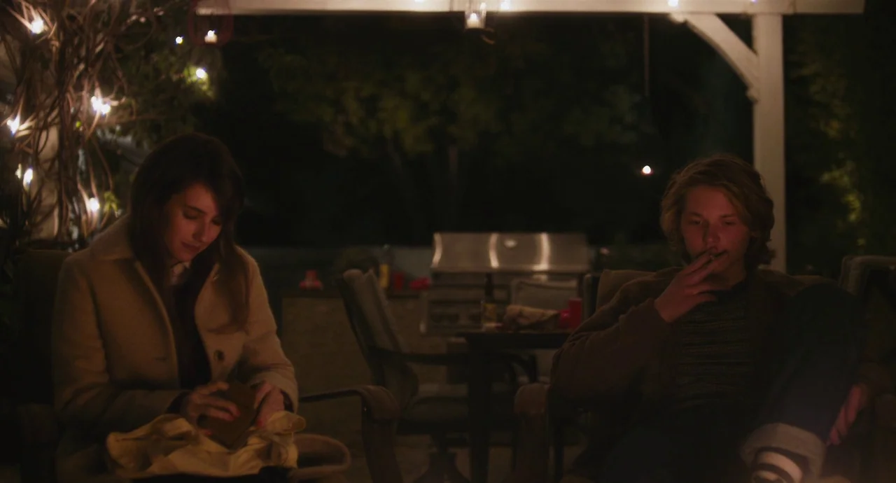 Palo Alto (2013) movie still: medium — A nighttime patio space features string lights draped above two seated individuals. A man smokes a cigarette…
