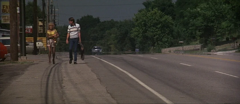 Nashville (1975) movie still: fullbody — A rural highway features a paved road bordered by a grassy verge and trees. A young man and woman walk…