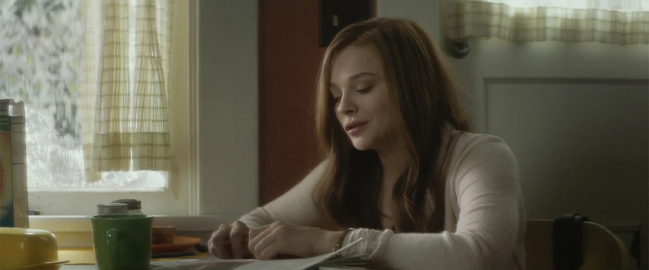 If I Stay (2014) movie still: medium — A bright domestic area features a wooden table near a window with sheer curtains. A young woman in a cream…
