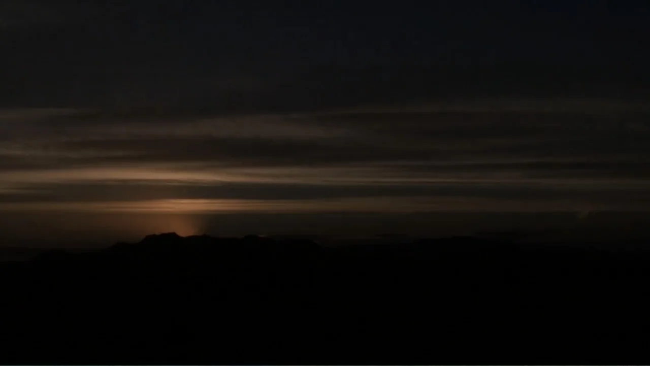Savageland (2015) movie still: establishing — A dark mountainous silhouette sits against a dim sunset sky with layered cloud formations. The horizon…