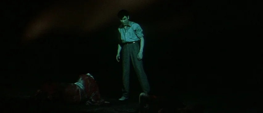 Jigoku (1960) movie still: wide — A dark, undefined space contains a single standing man and two figures lying prostrate. The standing man looks…
