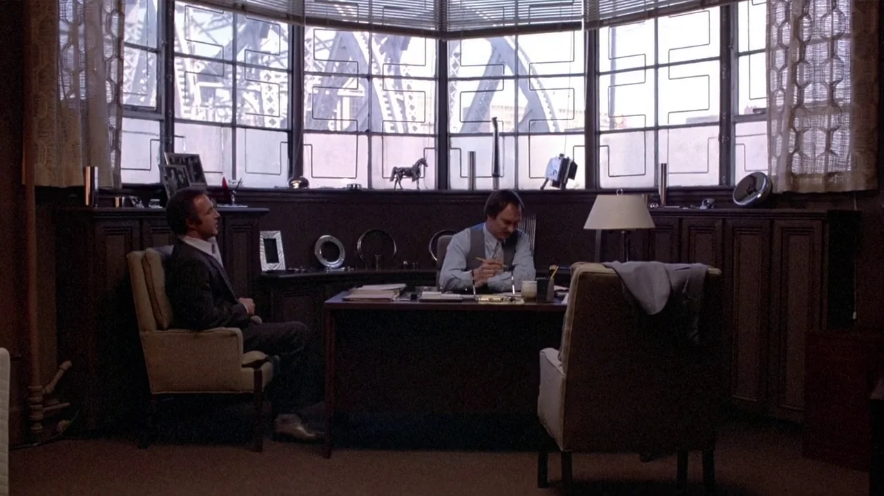 Thief (1981) movie still: wide — A wood-paneled room features a large window overlooking a bridge structure outside. Two men interact in the space…