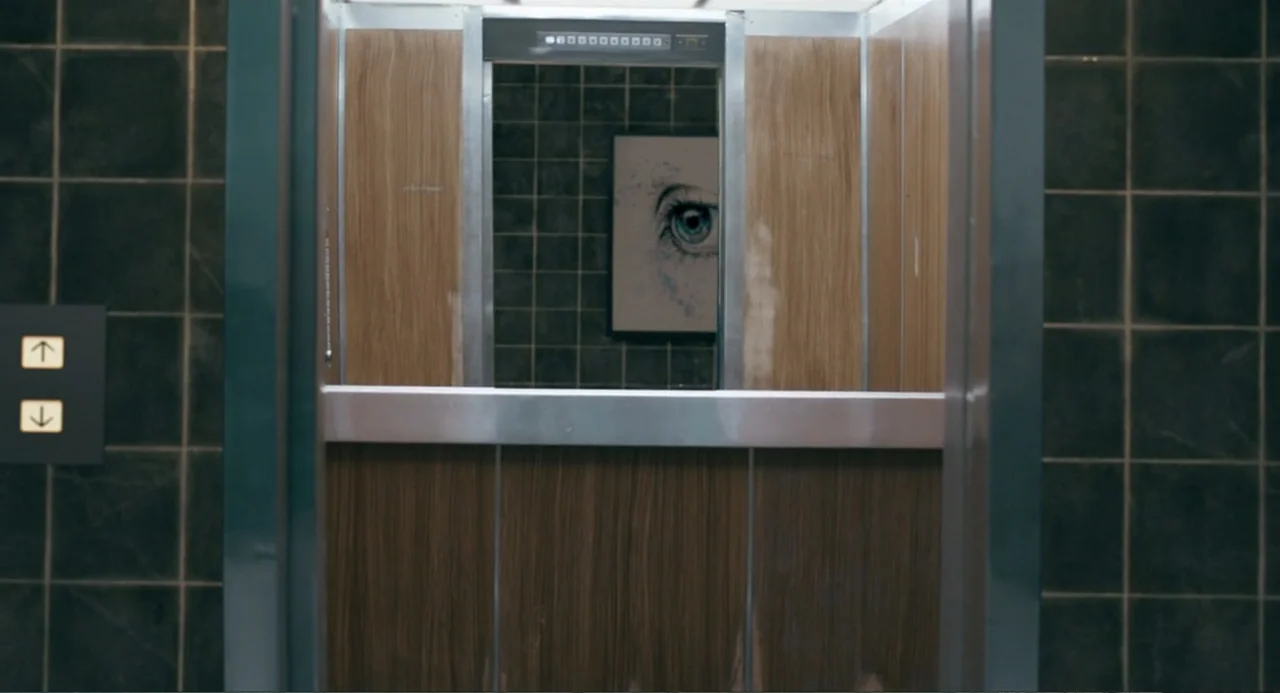 The Lift (2021) movie still: establishing — An elevator interior features wood-paneled walls and dark charcoal-colored tiles on the sides. A framed…