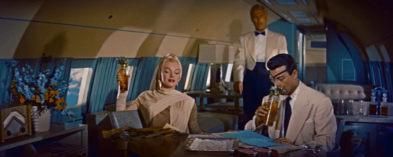 How to Marry a Millionaire movie still: wide — A luxury airplane cabin features blue curtains and curved walls with retro styling. A woman and two…