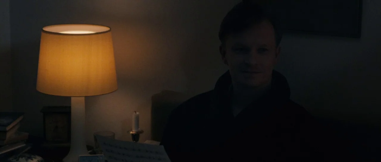 Sound of Noise (2010) movie still: medium — A dimly lit room features a table lamp casting a warm glow on a man. The man sits in darkness, holding a…