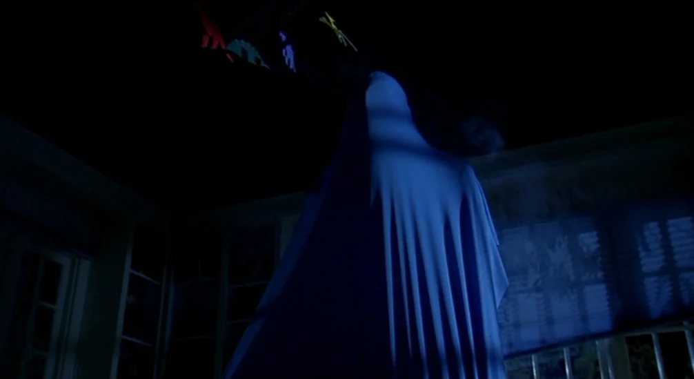 New Nightmare (1994) movie still: medium — A dark, dimly lit room contains a figure covered completely by a blue sheet. The figure stands centrally…