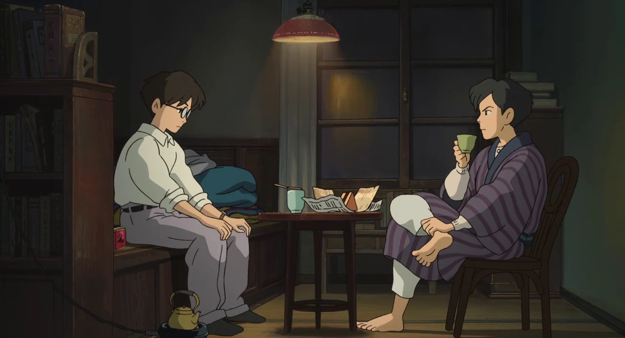 The Wind Rises (2013) movie still: medium — A dimly lit room contains wooden furniture, a bookshelf, and a window at night. Two young men sit facing…