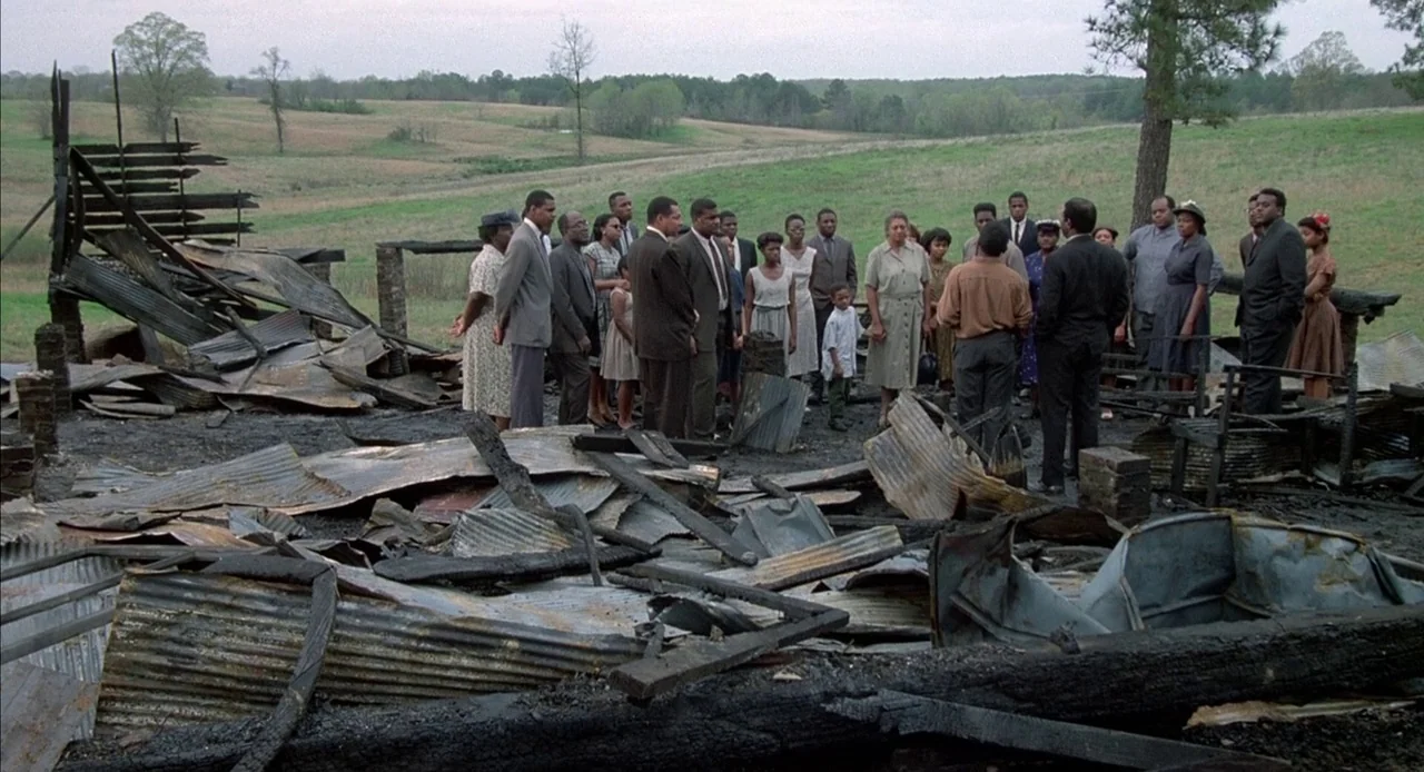Mississippi Burning (1988) movie still: wide — A group of African American people stand in a field surrounding burnt, debris-filled ruins. They are…