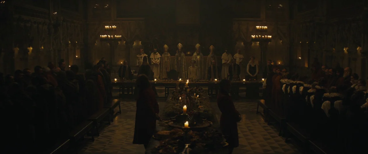 Macbeth (2015) movie still: wide — A long stone hall features rows of seated guests and a central banquet table. Figures stand along the back wall…