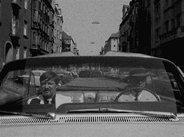 The American Soldier (1970) movie still: medium — A city street flanked by tall, older apartment buildings is viewed through a car windshield. Two…