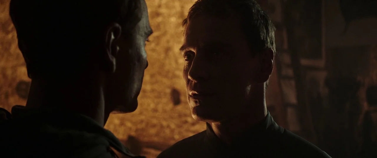 Alien: Covenant (2017) movie still: closeup — A dark indoor setting is lit by a single warm light source against stone. Two men stand face-to-face…