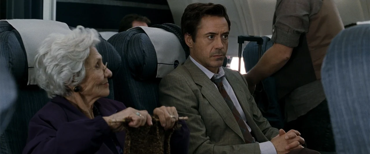 Due Date (2010) movie still: medium — An airplane cabin with rows of blue cushioned seats and white headrests. A man sits beside an elderly woman…