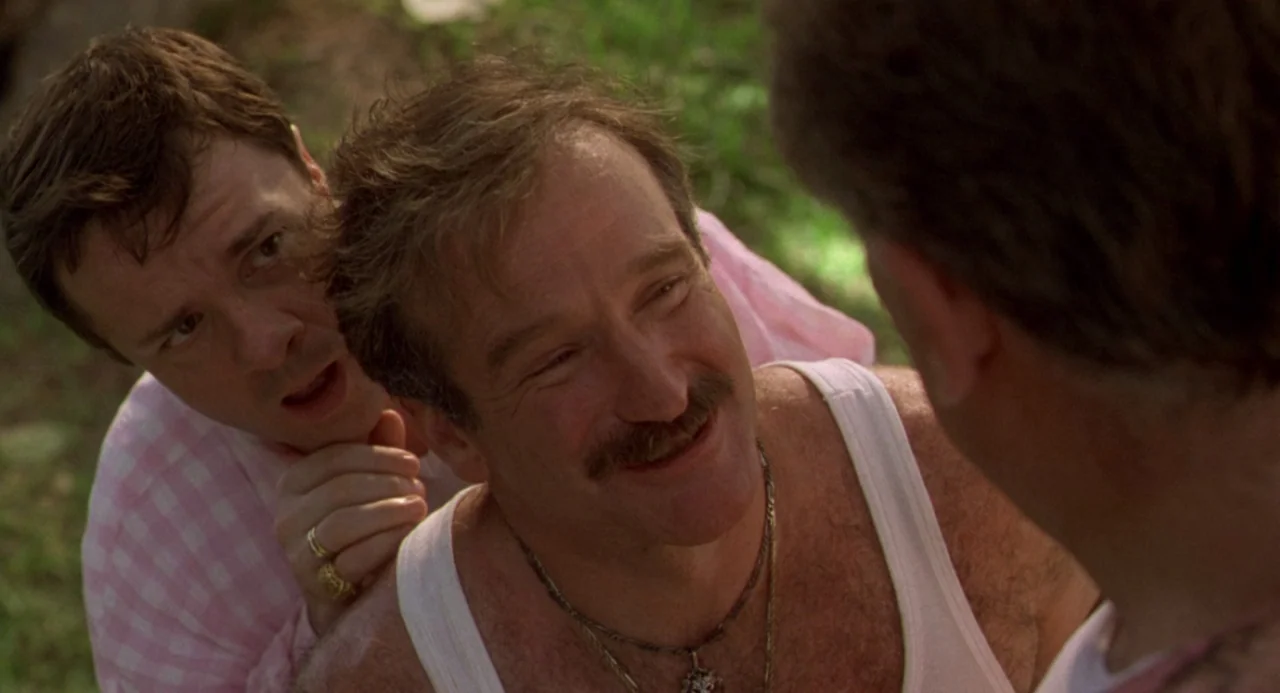 The Birdcage (1996) movie still: medium — Two men stand outdoors in front of a green grassy background. One man smiles at another person who is…