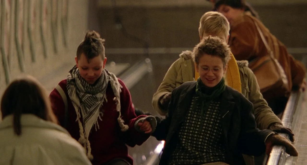 We Are The Best! (2013) movie still: medium — Two teenagers stand on an upward-moving escalator in a concrete public space. They are smiling at each…