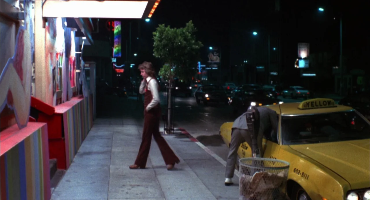 The Killing of a Chinese Bookie (1976) movie still: fullbody — A night city sidewalk features a yellow taxi parked next to a colorful building wall.…