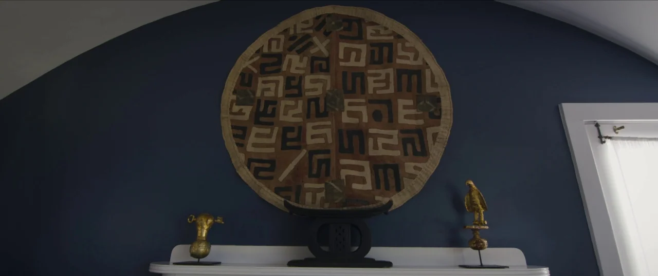Da Sweet Blood of Jesus (2014) movie still: medium — A circular African textile hangs on a matte dark blue wall above a mantel. Three decorative…