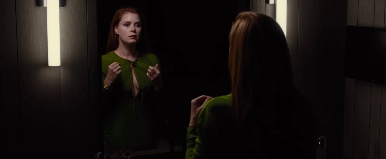 Nocturnal Animals (2016) movie still: medium — A woman stands in front of a mirror in a dark, wood-paneled dressing area. She adjusts the string tie…
