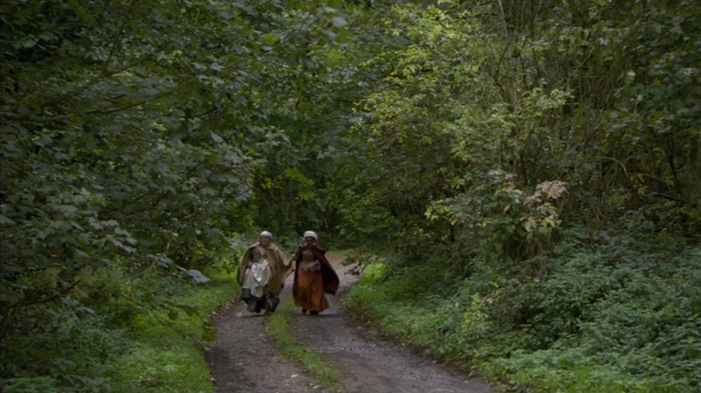 A Cock and Bull Story (2005) movie still: wide — A dirt track leads through a dense forest with thick green overhead foliage. Two women in…