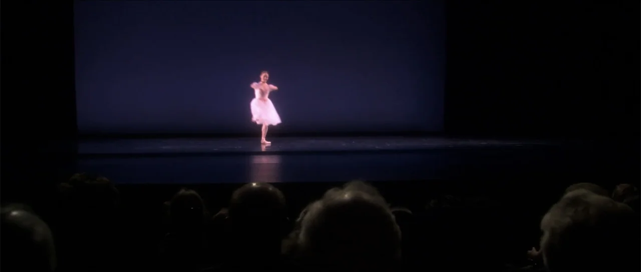 El acto (1989) movie still: wide — A ballet dancer performs on a dark theater stage under a bright spotlight. The audience sits in complete shadow…