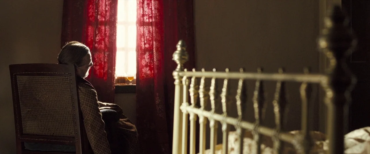 The Proposition (2005) movie still: medium — An interior space contains a wooden chair and a metal bed frame in foreground. An elderly man sits in…