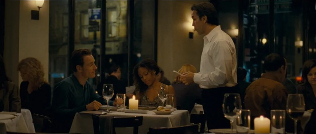 Shame (2011) movie still: medium — An upscale dining room features white tablecloths and dark evening lighting through large glass windows. A man…