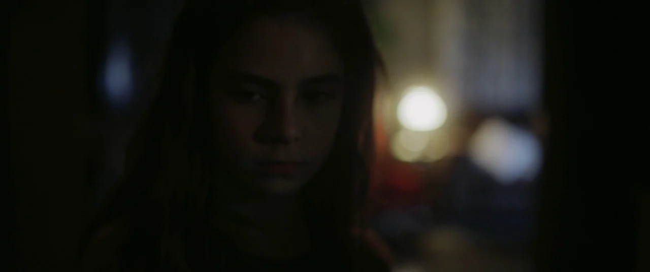 Mustang (2015) movie still: closeup — A dimly lit interior space features a person in the foreground. A soft-focus lamp glows in the blurred…