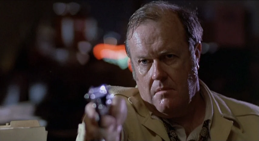 Blood Simple (1984) movie still: closeup — An interior space at night features an older man in a tan jacket. He holds a revolver pointed directly…