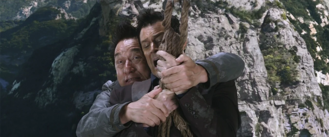 Skiptrace (2016) movie still: medium — A vertical rocky mountain side serves as a backdrop for two men. The men grasp a thick rope with both hands…