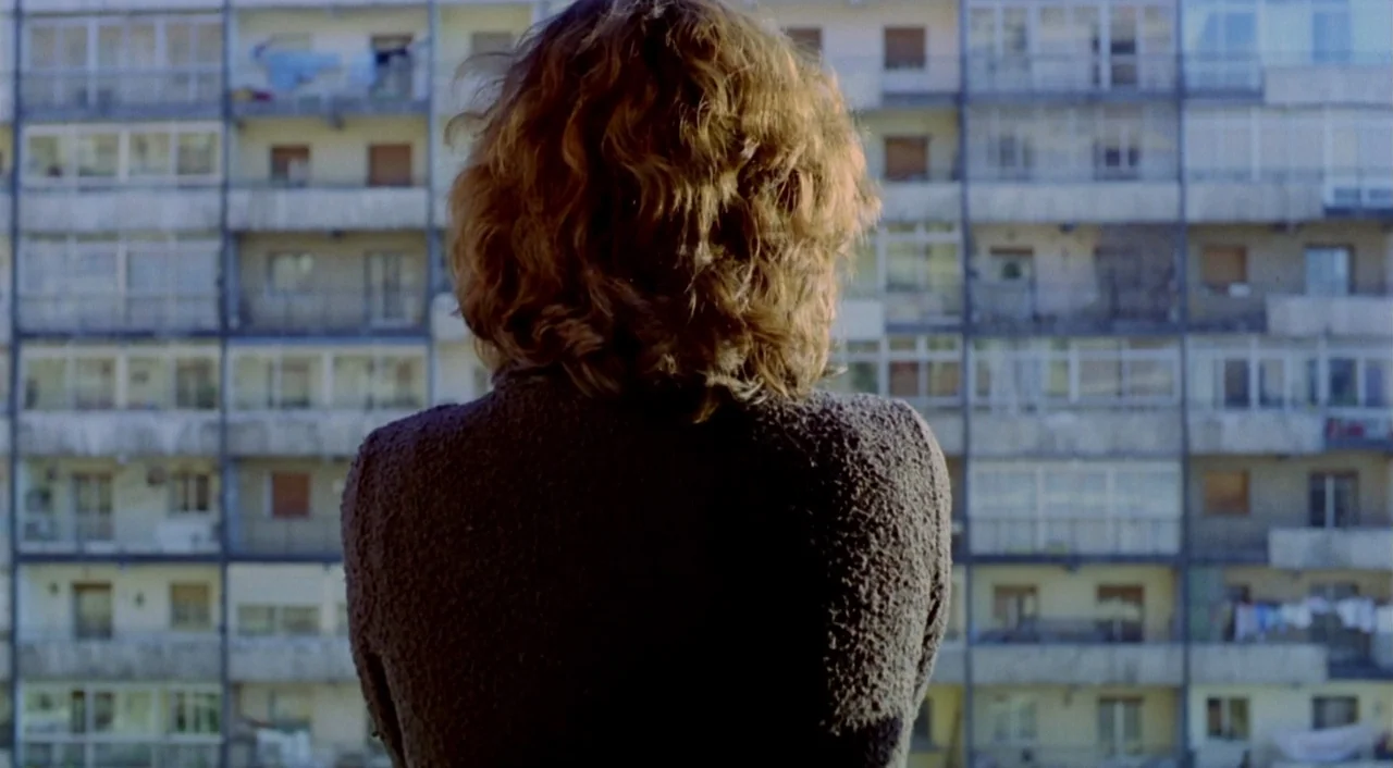 What Have I Done To Deserve This? (1984) movie still: medium — A woman stands in an interior room looking out toward a large concrete residential…