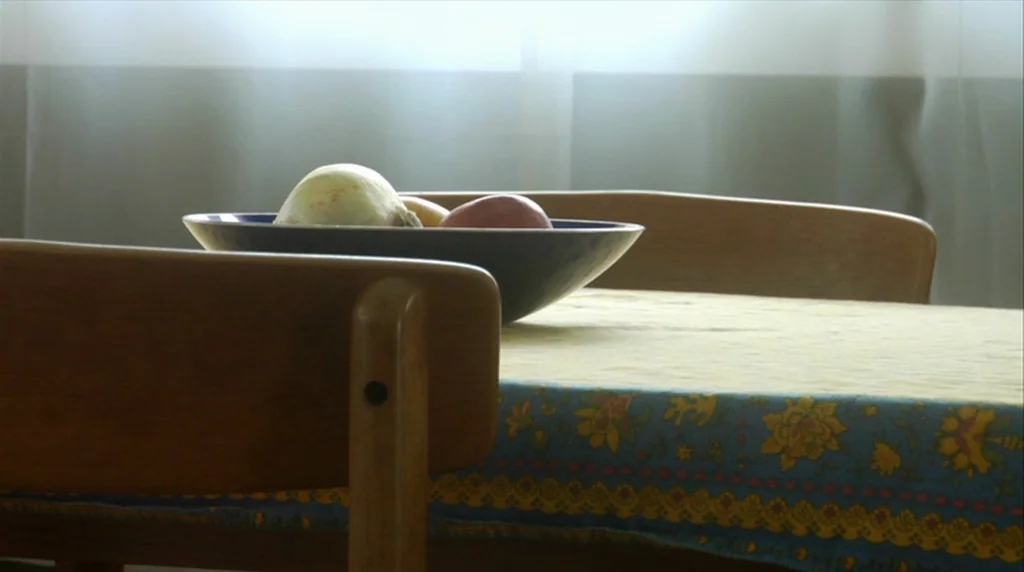 Humpday (2009) movie still: closeup — A wooden dining table is covered with a yellow and blue floral patterned tablecloth. A dark bowl containing an…