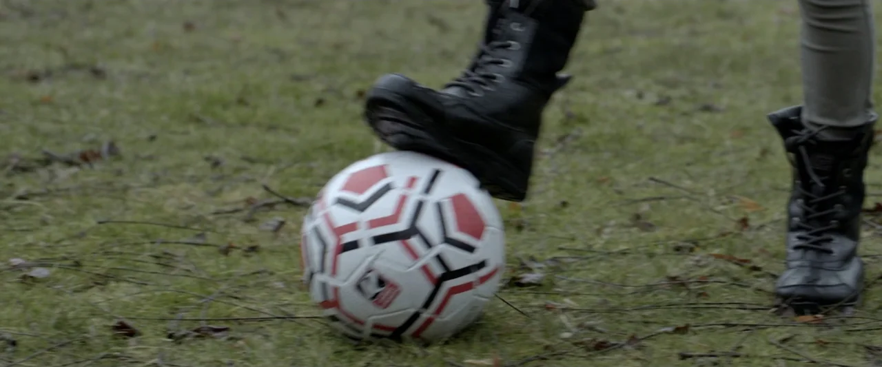 Motherly (2022) movie still: closeup — A soccer ball sits on sparse grass with twigs scattered across the ground. A person's boot rests on top of…