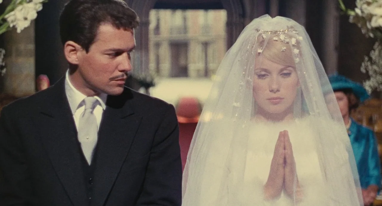 The Umbrellas Of Cherbourg (1964) movie still: medium — A stone interior space features a distant archway visible between two figures. The man in a…