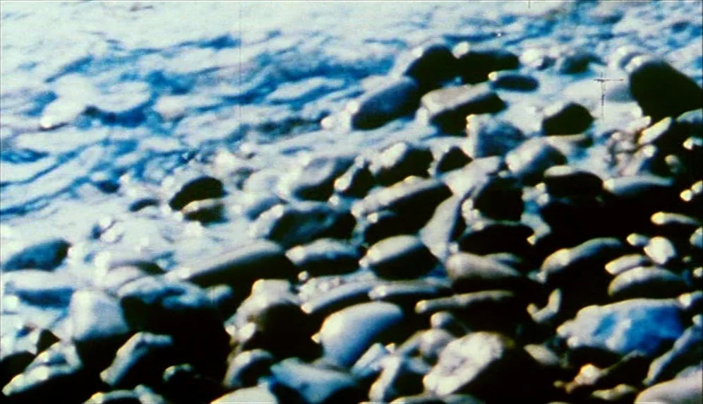 Nenette et Boni (1996) movie still: closeup — A close-up view of dark, smooth river pebbles resting on a shoreline. Shallow waves wash over the…