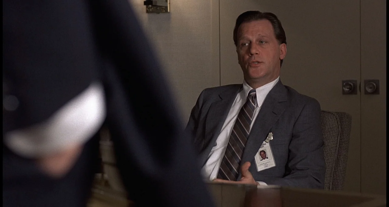 No Way Out (1987) movie still: medium — A professional 1990s office with light-colored walls and a desk in the foreground. A middle-aged man in a…