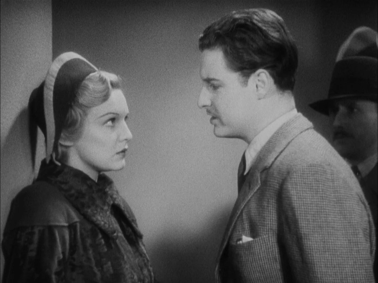 The 39 Steps (1935) movie still: medium — A plain interior wall serves as the backdrop for a conversation. A woman stands facing a man while another…